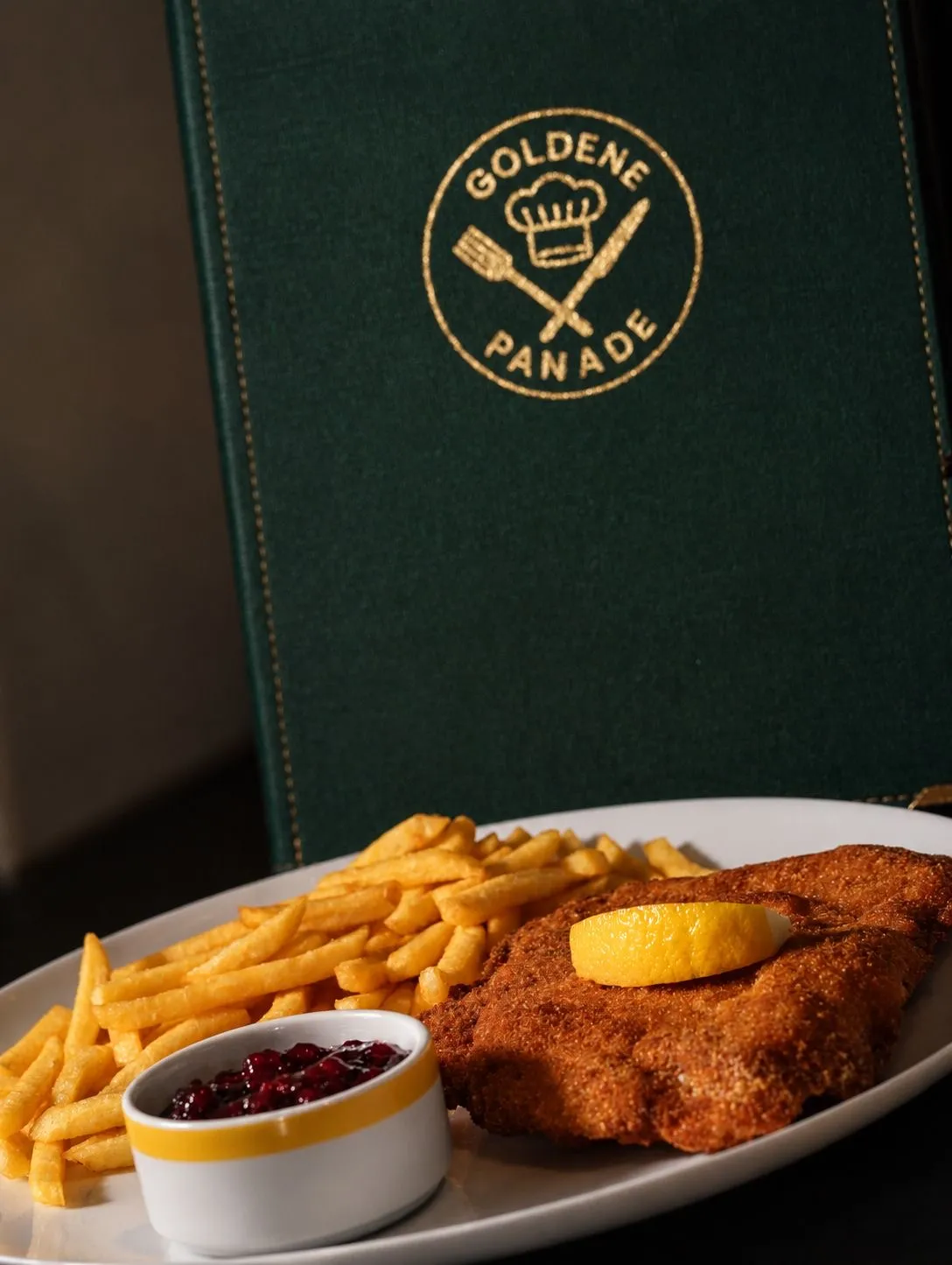 Grüne Speisekarte der Goldenen Panade neben einem Schnitzel mit Pommes und Preiselbeeren